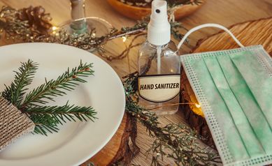 Christmas eve holiday party decorated table set with disposable medical mask and alcohol hand sanitizer bottle. Coronavirus (Covid 19) spreading prevention concept. Christmas micro led lights wire.