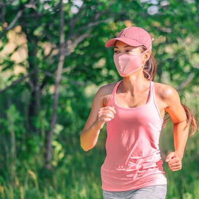 Mask wearing during exersice for COVID-19 protection Asian girl running outside with face covering while exercising jogging on run sport workout in summer park nature. Pink mask, cap, tank top